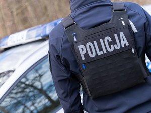Zdjęcie policjanta od storny pleców. Ubrany w kamizelkę taktyczną. W tle radiowóz policji.