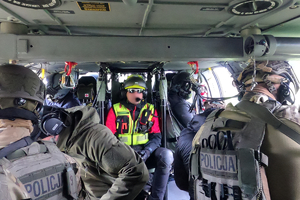 Widok wnętrza śmigłowca. Po lewej i prawej w burtach śmigłowca siedzący policjanci. Na wprost widoczna obsługa maszyny oraz uczestnik ćwiczeń w żółtej kamizelce.