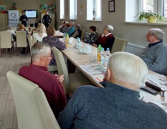 Zdjęcie wykonane w  Dziennym Domu Pomocy Caritas Diecezji Rzeszowskiej w Zagorzycach. W szerokiej perspektywie widać starsze osoby które siedzą przy stołach ustawionych wzdłuż pomieszczenia w kształcie prostokątnym. W dali widoczni są stojący dwaj funkcjonariusze policji. 