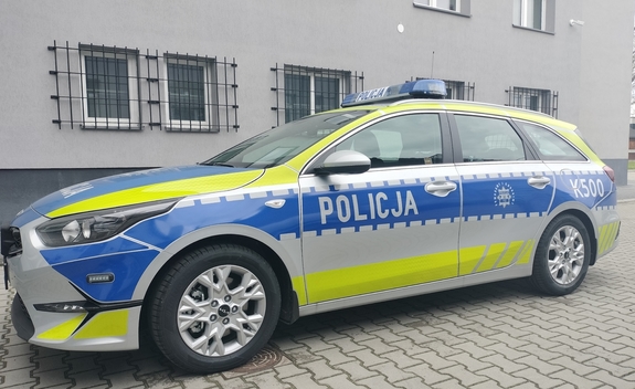 Na zdjęciu oznakowany  radiowóz policyjny marki KIA koloru srebrnego z logo Policji oraz nienaniesionymi na elementy radiowozu barwami koloru niebieskiego oraz seledynowego-neonowego.