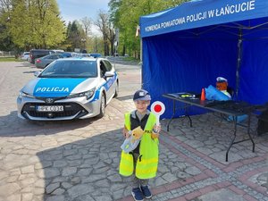 Chłopiec ubrany w policyjne umundurowanie pozuje przed policyjnym stoiskiem i oznakowanym radiowozem.