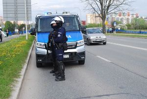 Na zdjęciu dwóch umundurowanych policjantów, za nimi radiowóz. Obok jadący samochód. W tle przechodnie i osoby jadące na rowerach