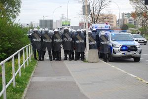 Na zdjęciu policjanci w pododdziale zwartym, ubrani w zestaw przeciwuderzeniowy, obok radiowóz