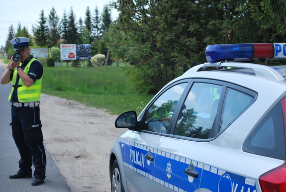 Na zdjęciu umundurowany policjant ruchu drogowego z miernikiem prędkości, mierzy prędkość pojazdów. Za nim radiowóz. W tle drzewa.