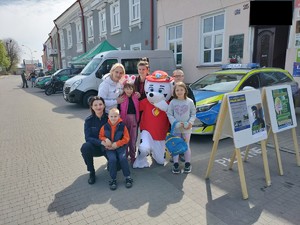 Na zdjęciu Policjantka w mundurze służbowym koloru granatowego z uczestnikami pikniku. W tle oznakowany radiowóz.