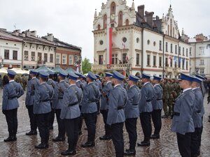 na zdjęciu stojący w szyku policjanci kompanii honorowej, zdjęcie wykonane z tyłu, w tle budynki