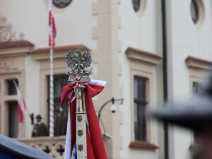 wyostrzona na zdjęciu głowica sztandaru z orłem w koronie i biało-czerwoną kokardą, w tle rozmyty budynek z masztem i biało-czerwoną flagą