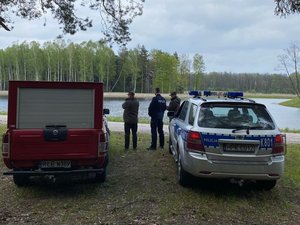 Policjant wraz z dwoma strażnikami leśnymi pełni służbę na terenie leśnym, w pobliżu stawu.
