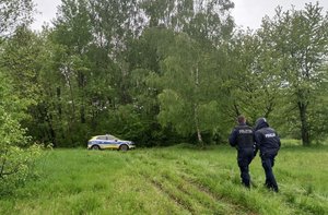 Na zdjęciu dwóch umundurowanych policjantów idących w kierunku lasu. W tle radiowóz policyjny i las.