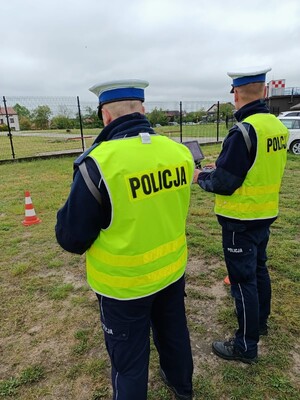 Zdjęcie przedstawia funkcjonariuszy Policji którzy sterują dronem elektronicznie. Ubrani w granatowe mundury z napisem Policja oraz zółte odblaskowe kamizelki.