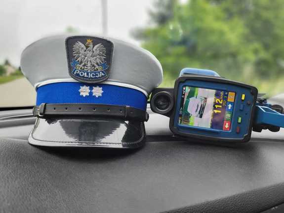 Na zdjęciu czapka służbowa policjanta w stopniu aspiranta ułożona na podszybiu pojazdu, obok ręczny miernik prędkości z utrwalona prędkością 112 km/h.