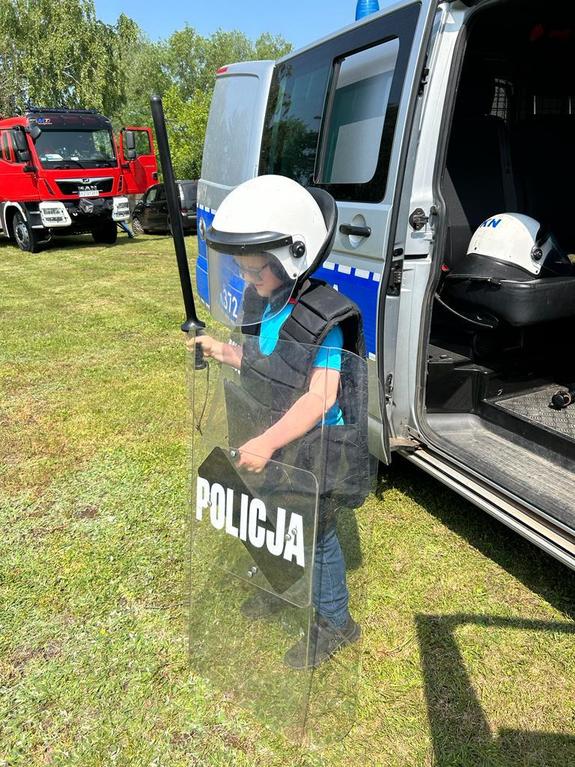 na trawniku stoi radiowóz typu bus przed radiowozem stoi dziecko ubrane w kamizelkę przeciwuderzeniową w ręku trzyma pałkę szturmową na głowie ubrany biały kask w drugiej ręce przezroczysta tarcza z napisem policja w tle wóz strażacki
