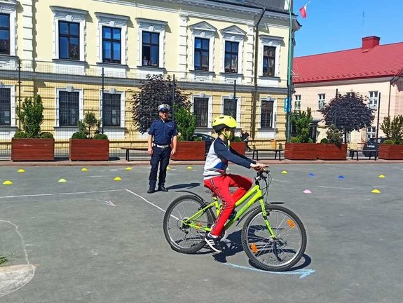 Po torze przygotowanym na boisku szkolnym rowerem jedzie chłopiec. Obok stoi oceniający jazdę rowerzysty policjant.