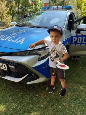 Chłopiec w czapce policyjnej trzymający tarczę do zatrzymywania pojazdów na tle radiowozu.