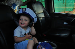 Mały chłopiec siedzący w radiowozie. Na głowie ma za dużą czapkę policjanta ruchu drogowego, uśmiecha się, a w ręce trzyma mikrofon do stacji radiowej