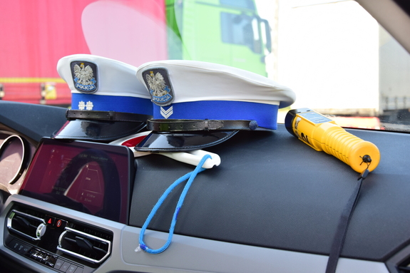 Deska rozdzielcza policyjnego radiowozu. Na niej leżą dwie czapki policjantów ruchu drogowego oraz żółte urządzenie do sprawdzania obecności alkoholu w wydychanym powietrzu