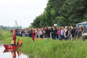 Na zdjęciu uczestnicy spotkania stoją przy akwenie wodnym, na wodzie strażacy w łódce.