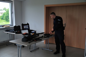 Policjant stojący przy stole, na którym znajduje się kilka jednostek broni, hełmy oraz alkomat i ręczny miernik prędkości.