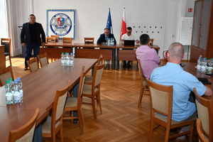 Szkolenie w auli Komendy Wojewódzkiej Policji w Rzeszowie. Prelegenci siedzą za stołem, przed sobą mają laptopy, za nimi flaga Polski i Unii Europejskiej oraz policyjna gwiazda z herbem Podkarpacia i napisem Komenda Wojewódzka Policji w Rzeszowie