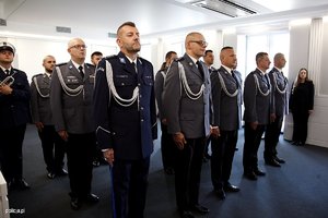 Zdjęcie przedstawia wnętrze sali, w której ustawieni są pracownicy Policji, umundurowani policjanci, poczet sztandarowy. Policjanci i pracownicy odbierają odznaczenia od Komendanta.