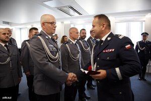 Zdjęcie przedstawia wnętrze sali, w której ustawieni są pracownicy Policji, umundurowani policjanci, poczet sztandarowy. Policjanci i pracownicy odbierają odznaczenia od Komendanta.