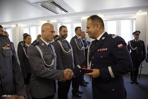 Zdjęcie przedstawia wnętrze sali, w której ustawieni są pracownicy Policji, umundurowani policjanci, poczet sztandarowy. Policjanci i pracownicy odbierają odznaczenia od Komendanta.