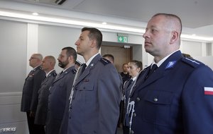 Zdjęcie przedstawia wnętrze sali, w której ustawieni są pracownicy Policji, umundurowani policjanci, poczet sztandarowy. Policjanci i pracownicy odbierają odznaczenia od Komendanta.