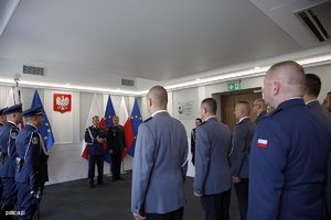 Zdjęcie przedstawia wnętrze sali, w której ustawieni są pracownicy Policji, umundurowani policjanci, poczet sztandarowy. Policjanci i pracownicy odbierają odznaczenia od Komendanta.