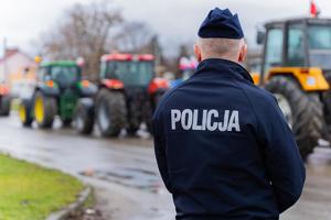 Na pierwszym planie umundurowany policjant, odwrócony plecami do obiektywu. W tle widać rząd ciągników rolniczych