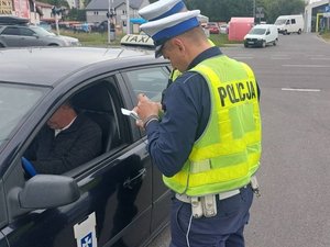 umundurowani policjanci ruchu drogowego w żółtych kamizelkach z napisem Policja, stoją przy osobowym samochodzie TAXI, kontrolują kierującego.