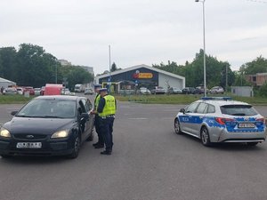 umundurowani policjanci ruchu drogowego w żółtych kamizelkach z napisem Policja, stoją przy osobowym samochodzie TAXI, kontrolują kierującego. Z prawej strony ustawiony radiowóz.