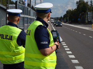 na zdjęciu dwaj umundurowani policjanci ruchu drogowego, w żółtych kamizelkach stojący przy ruchliwej drodze z urządzeniem do pomiaru prędkości