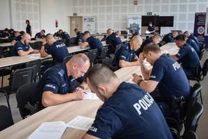 Policjanci podczas zmagań w konkursie na najlepszą parę patrolową. Na zdjęciu policjanci biorący udział w konkursie podczas pisania testu wiedzy, siedzą przy stołach.