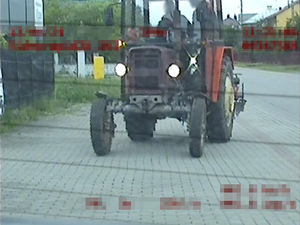 Na zdjęciu widoczny traktor a w środku dwie osoby. Traktor jedzie drogą, ma włączone światła.