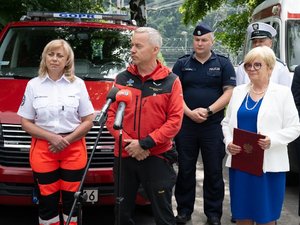 Wojewoda Podkarpacki, przedstawiciele służb, inspekcji i straży ustawieni na tle pojazdów służbowych podczas briefingu udzielają informacji mediom.