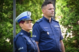 Dwoje policjantów i policjantka stoją przy mikrofonach podczas briefingu. W tle zielone krzewy i drzewa.
