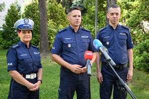 Dwoje policjantów i policjantka stoją przy mikrofonach podczas briefingu. W tle zielone krzewy i drzewa.