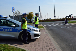 Po lewej stronie policjant i policjantka stoją przy radiowozie. W tle po prawej osoby przechodzące przez oznakowane przejście dla pieszych