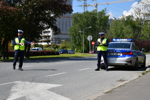 Policjanci ruchu drogowego stoją przy radiowozie. Policjant po lewej wskazuje nadjeżdżającemu pojazdowi miejsce do zatrzymania