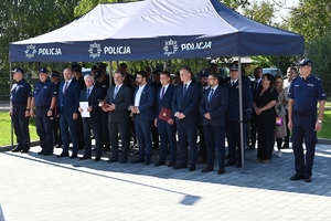 Policjanci i zaproszeni goście podczas uroczystości otwarcia komisariatu. Policjanci stoją na parkingu przed budynkiem.