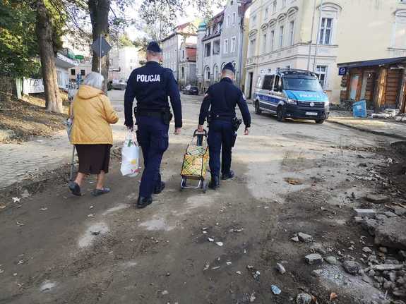 dwaj umundurowani policjanci z seniorką, pomagają jej nieść produkty żywnościowe