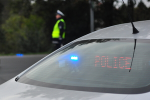Na pierwszym planie szyba tylnego radiowozu z sygnałem świetlnym i napisem POLICE. W tle policjant ruchu drogowego w kamizelce odblaskowej.