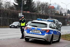 Policjantka mierząca prędkość z jaką poruszają się kierowcy jadący przez jedną z rzeszowskich ulic. Obok niej stoi oznakowany radiowóz. w Tle inne pojazdy.