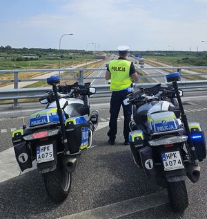 Policjant ruchu drogowego stojący na wiadukcie i sprawdzający odległość między pojazdami, na pierwszym planie dwa motocykle służbowe