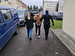 Policjantka z opaską POLICJA z lewej i policjant z prawej prowadzą zatrzymanego (w środku)