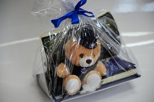 Policyjne gadżety przekazane na licytację. Voucher, policyjna maskotka, kalendarz, notatnik, proporczyk, kubek, smycz - w postaci paczki opakowanej w folię z niebieską wstążką.