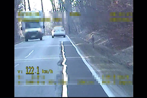Widoczny samochód jadący przed radiowozem, z lewej strony widać przejeżdżające z przeciwka auta. W dolnym lewym rogu pojawia się żółty napis 122.1 km/h