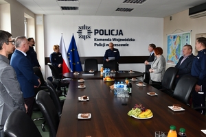 Uczestnicy spotkania w sali sztabowej Komendy Wojewódzkiej Policji w Rzeszowie. Uczestnicy siedzą przy stole, w tle na ścianie logo Komendy Wojewódzkiej Policji w Rzeszowie i napis  Komenda Wojewódzka Policji w Rzeszowie.