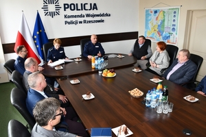 Uczestnicy spotkania w sali sztabowej Komendy Wojewódzkiej Policji w Rzeszowie. Uczestnicy siedzą przy stole, w tle na ścianie logo Komendy Wojewódzkiej Policji w Rzeszowie i napis  Komenda Wojewódzka Policji w Rzeszowie.
