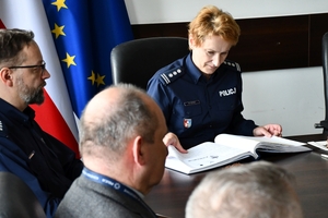 Uczestnicy spotkania w sali sztabowej Komendy Wojewódzkiej Policji w Rzeszowie. Uczestnicy siedzą przy stole, w tle na ścianie logo Komendy Wojewódzkiej Policji w Rzeszowie i napis  Komenda Wojewódzka Policji w Rzeszowie.